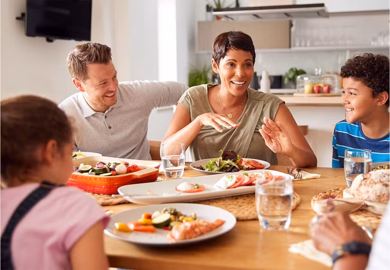 A family of four sits around a dinner table, smiling and talking while enjoying a meal together. Plates of food and glasses of water are on the table in a bright, modern dining area.