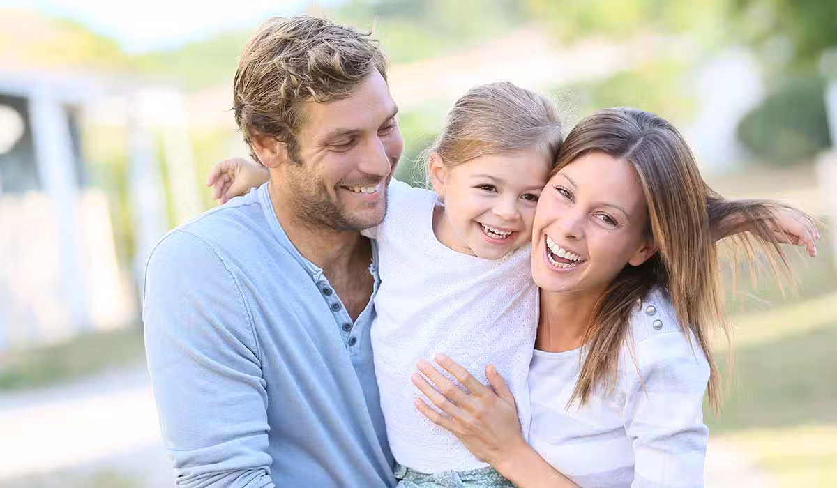 A happy family of three‚Äîan adult man, an adult woman, and a young girl‚Äîsmiles and embraces each other outdoors on a sunny day. The girl is between the adults, who both look joyful and relaxed.