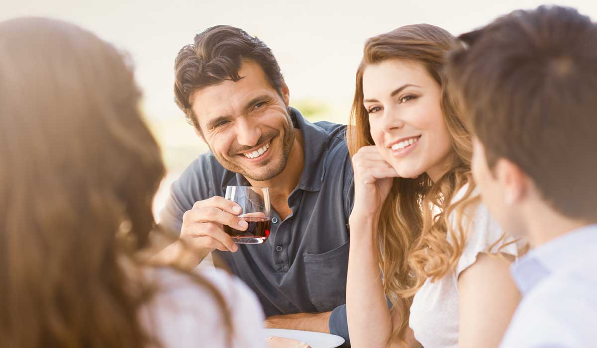 A group of four people sit together outdoors, smiling and talking. One man holds a glass of red wine. The atmosphere appears relaxed and friendly, with sunlight illuminating their faces.
