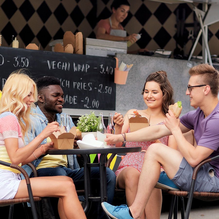 Four young adults sit at an outdoor table, eating takeout food and chatting cheerfully. A food stand with a chalkboard menu and a server in the background can be seen. Bottled drinks and plants are on the table.
