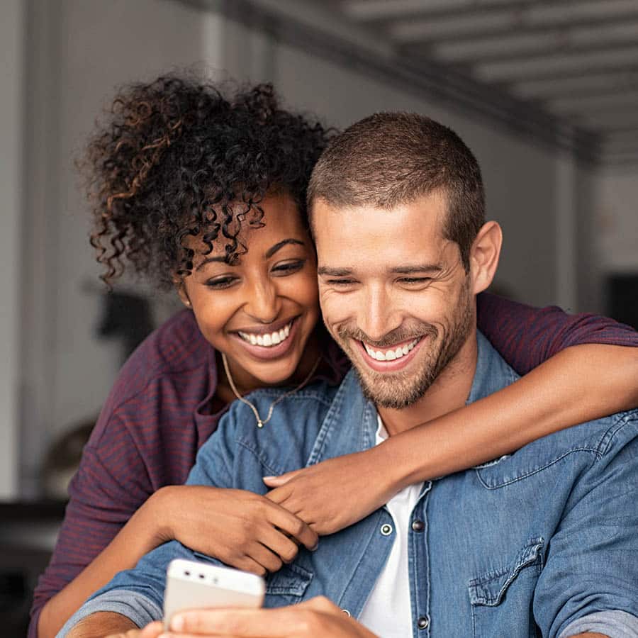 A smiling couple sits closely together, with the woman hugging the man from behind as they both look at a smartphone in his hand. They appear happy and relaxed in a casual indoor setting.