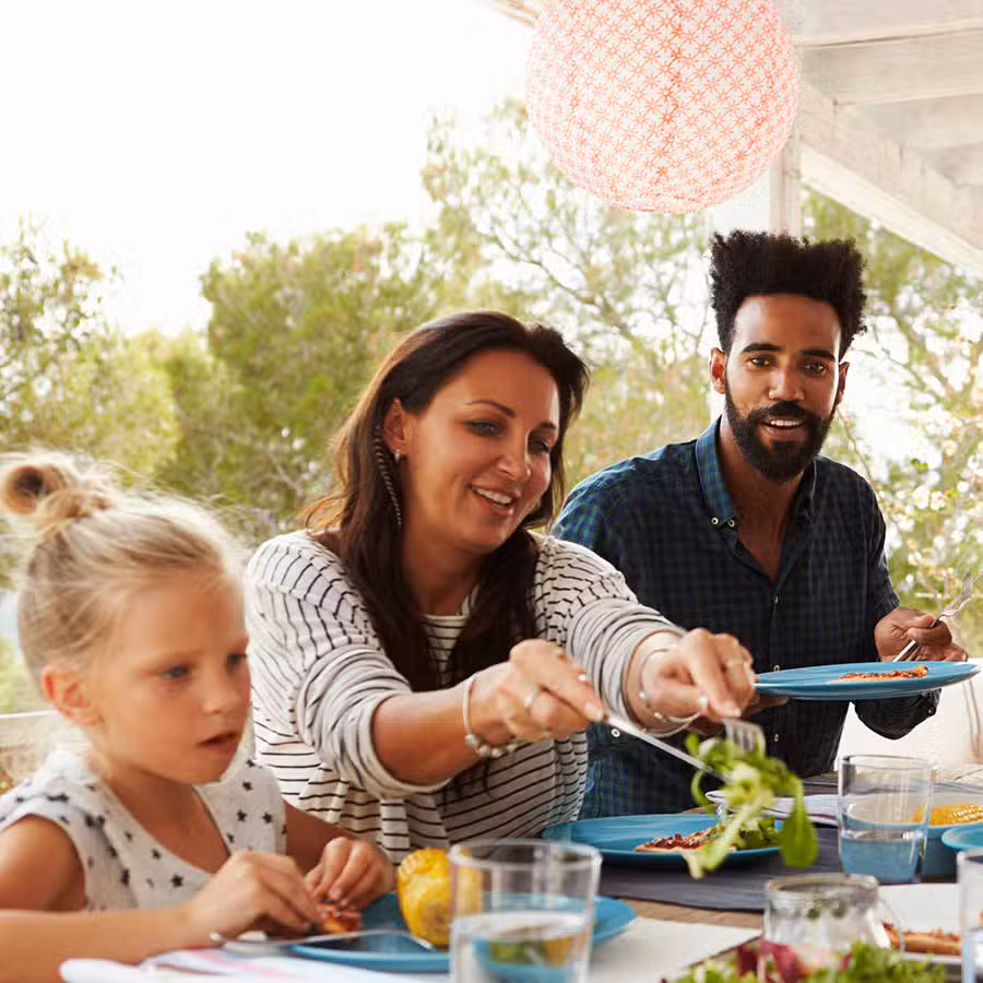Three people, two adults and a child, are sitting at a table outdoors, enjoying a meal together. The woman is serving salad, while the others are smiling. Trees and a pink lantern are visible in the background.