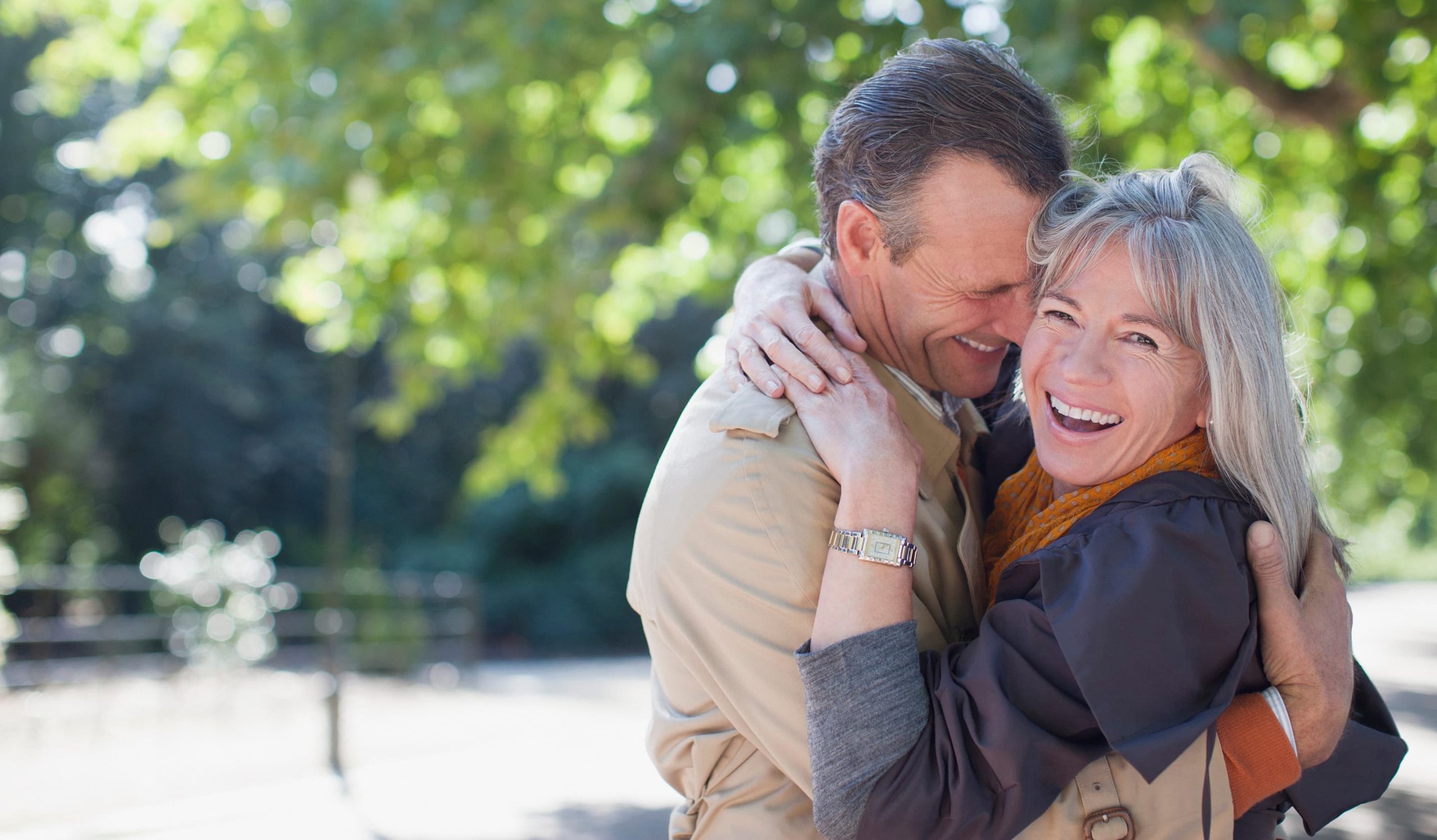 An older couple embraces and smiles joyfully outdoors, surrounded by trees and sunlight. The woman wears a gray jacket and orange scarf, while the man wears a beige coat. Both appear happy and content.